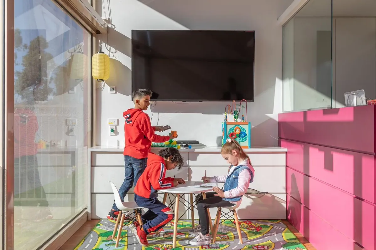 Three young children drawing together at a small table, with toys and an activity cube nearby.