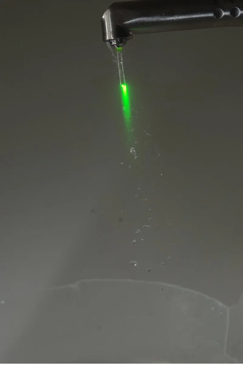Real photograph of the Fotona dental laser handpiece emitting its green aiming beam through cascading water droplets — visually demonstrating how the Er:YAG wavelength is absorbed by water, the principle that suits it to delicate soft-tissue procedures like frenectomy